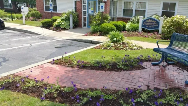 A blue bench sits next to a brick walkway surrounded by purple flowers and greenery in front of an apartment building.