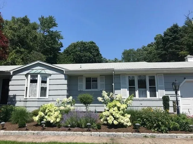 A light blue ranch house with white shutters and a flower bed against a bright blue sky.