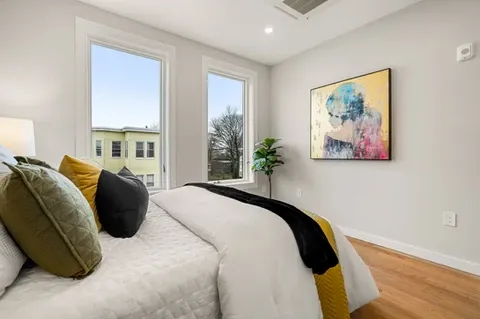 Bedroom on third floor with two windows, letting in ample light. Fully made bed set in the center of the room. A piece of art hanging  directly across from the bed.