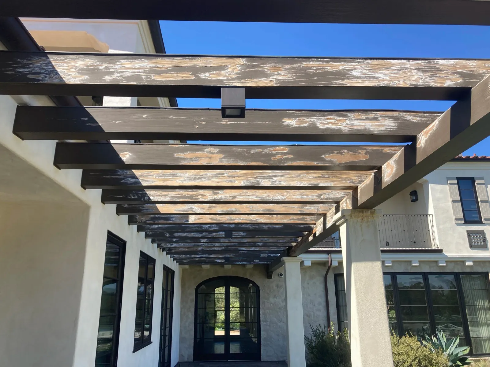 Wooden pergola with weathered planks over entrance to a modern house with black-framed windows and white stucco walls, blue sky in background.
