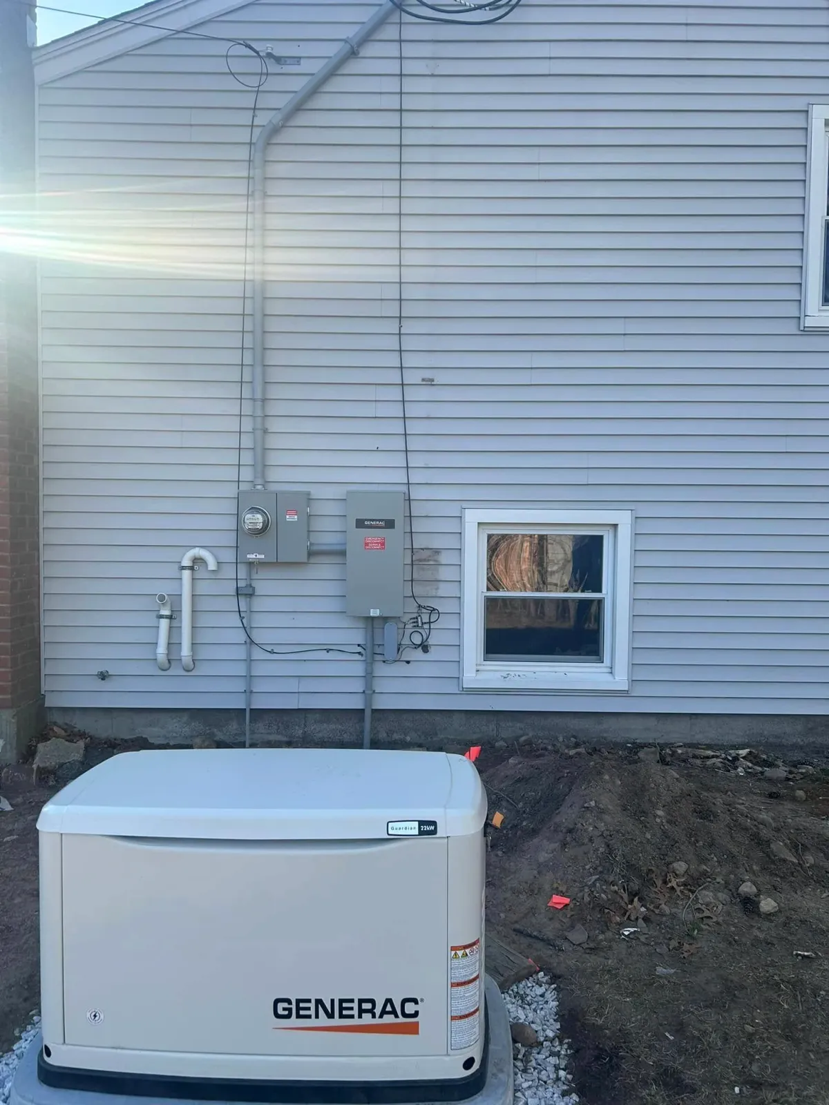 A Generac generator installed outside a house with gray siding, electrical boxes, and conduit pipes visible.