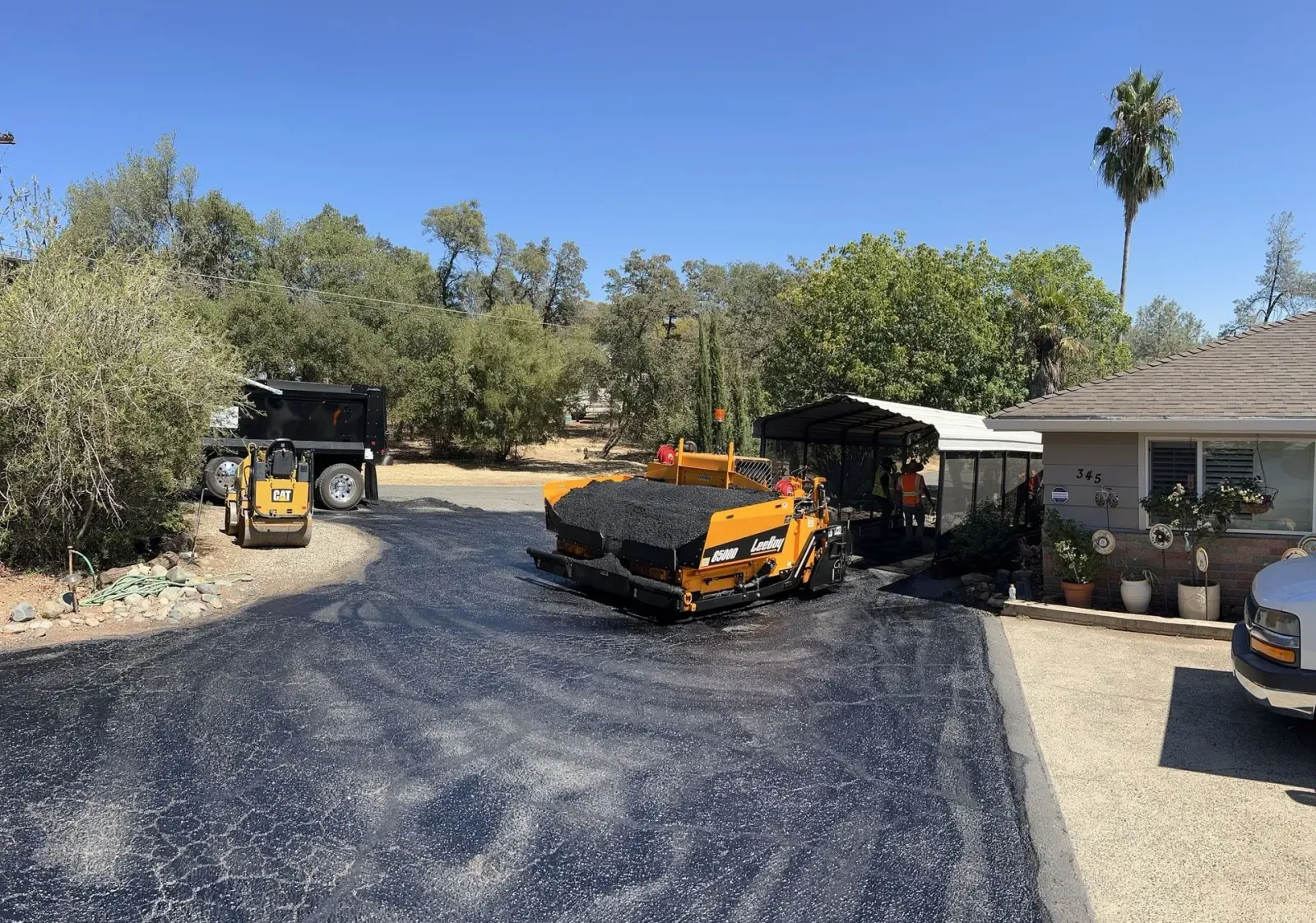Asphalt paving driveway. A large machine is spreading asphalt with workers.  Trees and house in background.