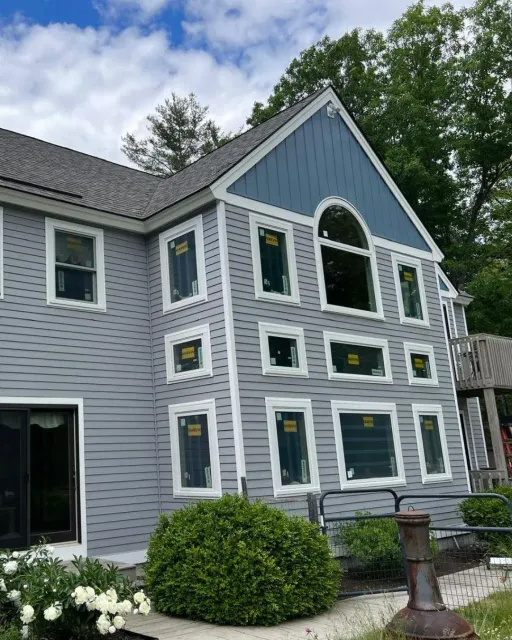 Full-house Marvin window replacement on a blue-grey clapboard home with a board-and-batten gable and an arched window, by Maia Construction