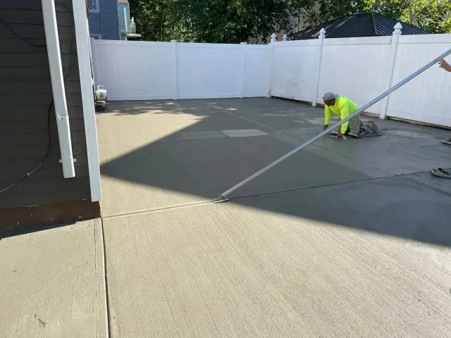Worker in neon safety gear smoothing fresh concrete in a fenced backyard.