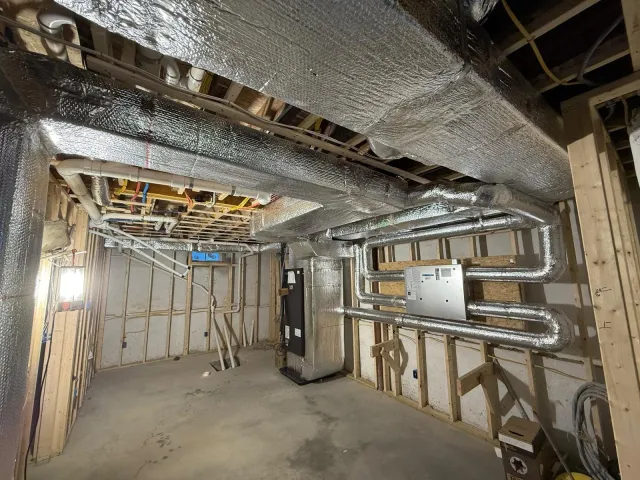 Construction site with exposed wooden framing and ductwork, including metal pipes, insulation, and electrical wiring.