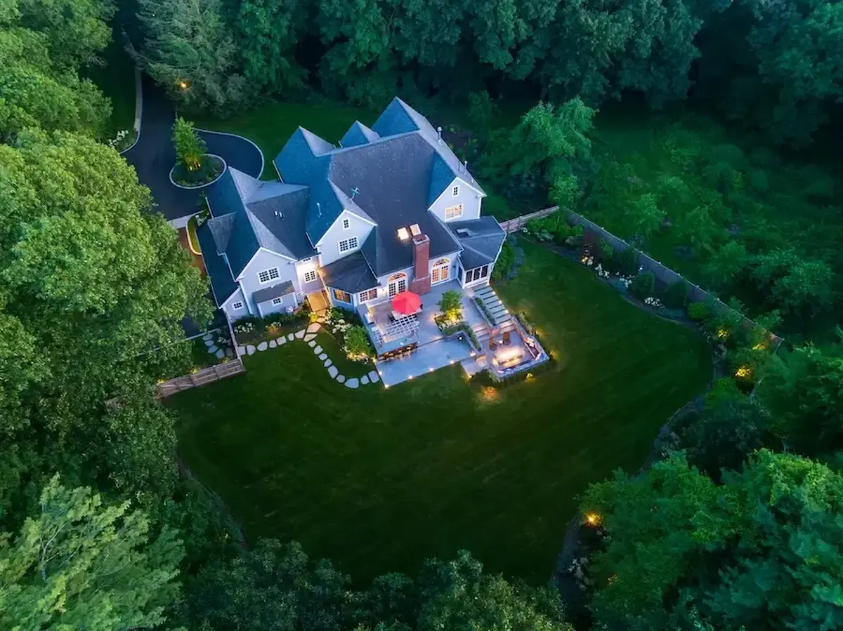 Aerial view of a large multi-wing gray house with a well-kept lawn, surrounded by dense trees and a curved stone path nearby.