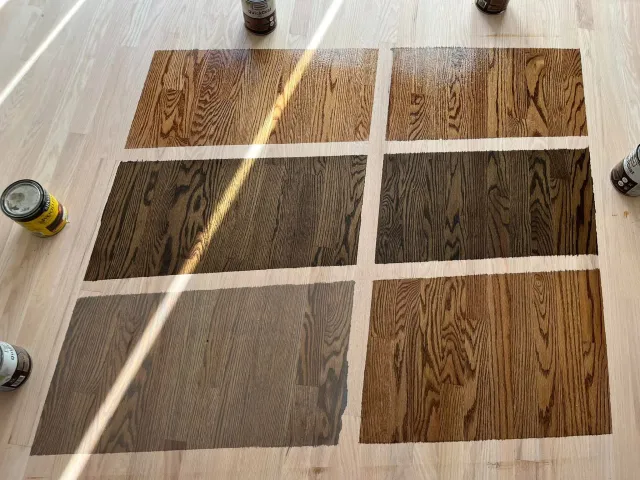 Sheet of wood flooring samples placed on a light wood floor, with cans of wood stain or finish surrounding the samples.