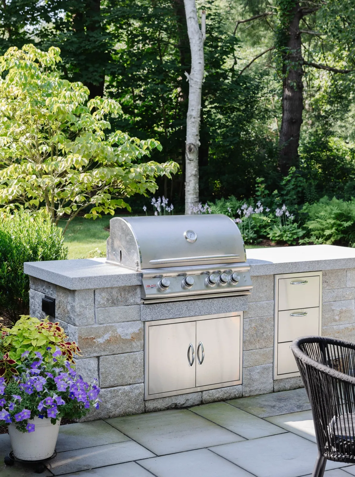 Outdoor kitchen with a built-in stainless steel grill, stone countertop, and storage cabinets, surrounded by plants and trees in a garden setting.