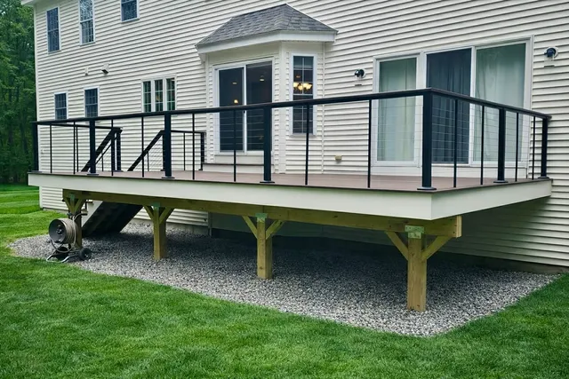 Raised wooden deck with black railing and stairs beside a beige house over gravel and grass