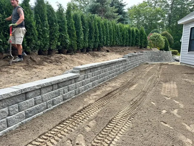 A landscaper trims hedges next to a retaining wall and dirt, near a house.