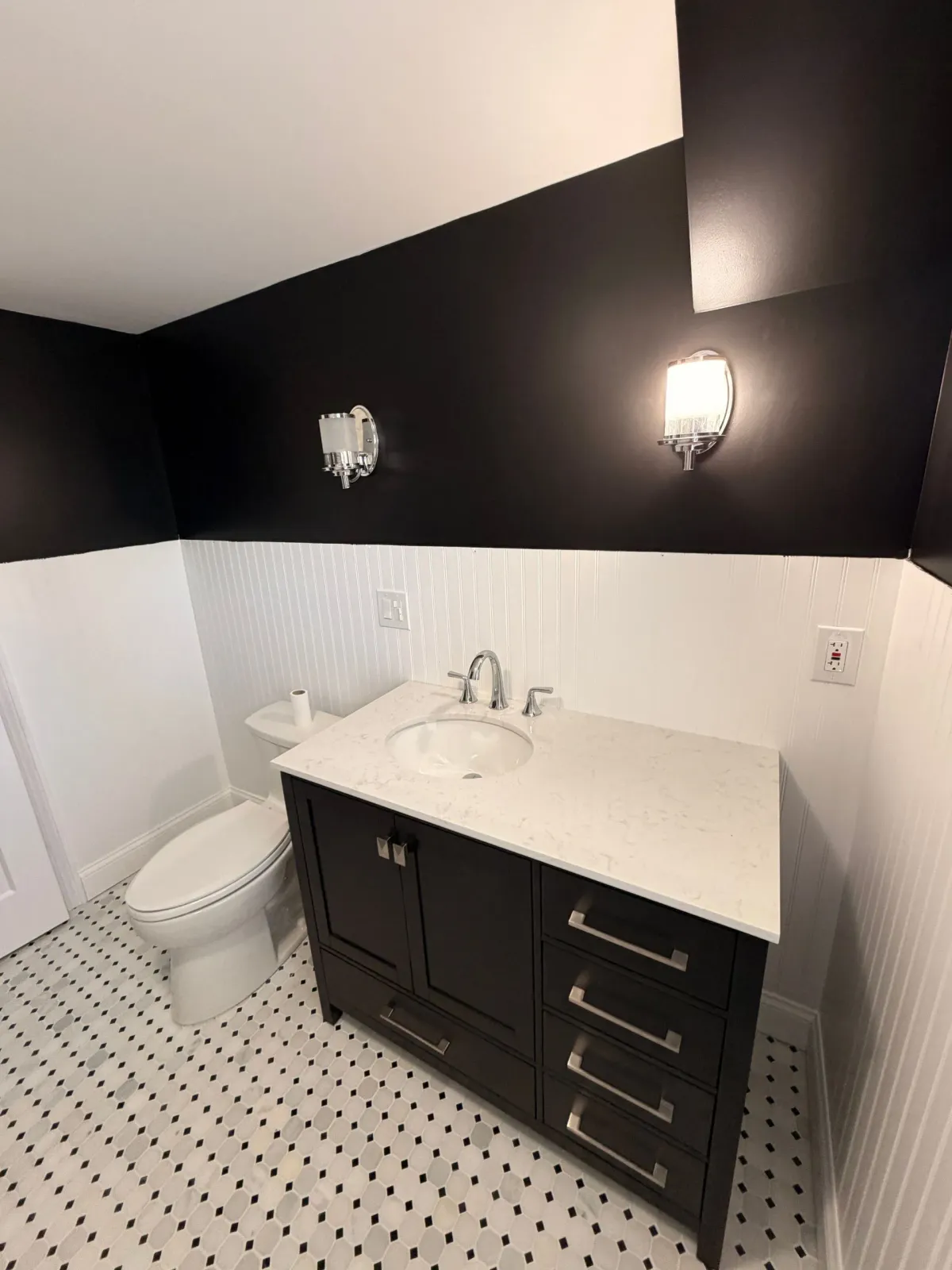 Black-and-white bathroom with pedestal sink, dark vanity, toilet, and patterned tile floor.