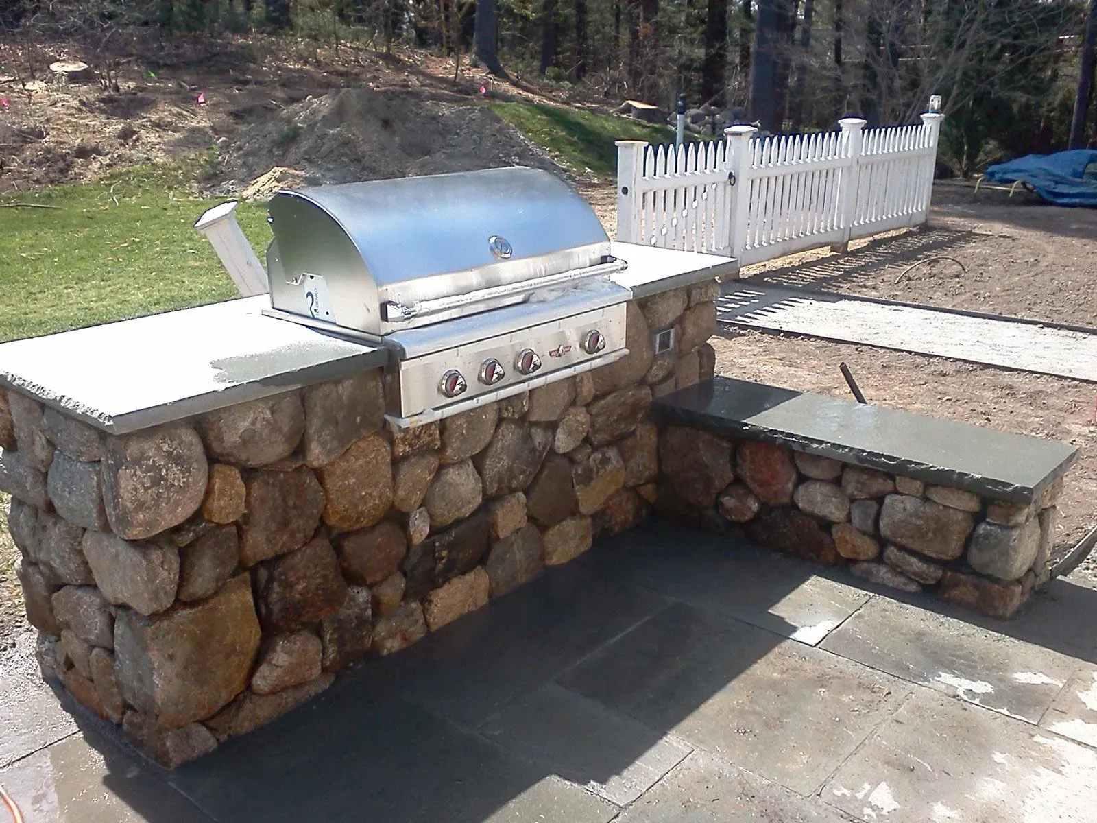 Stone outdoor patio with a stainless steel grill and built-in stone seating by a fenced yard