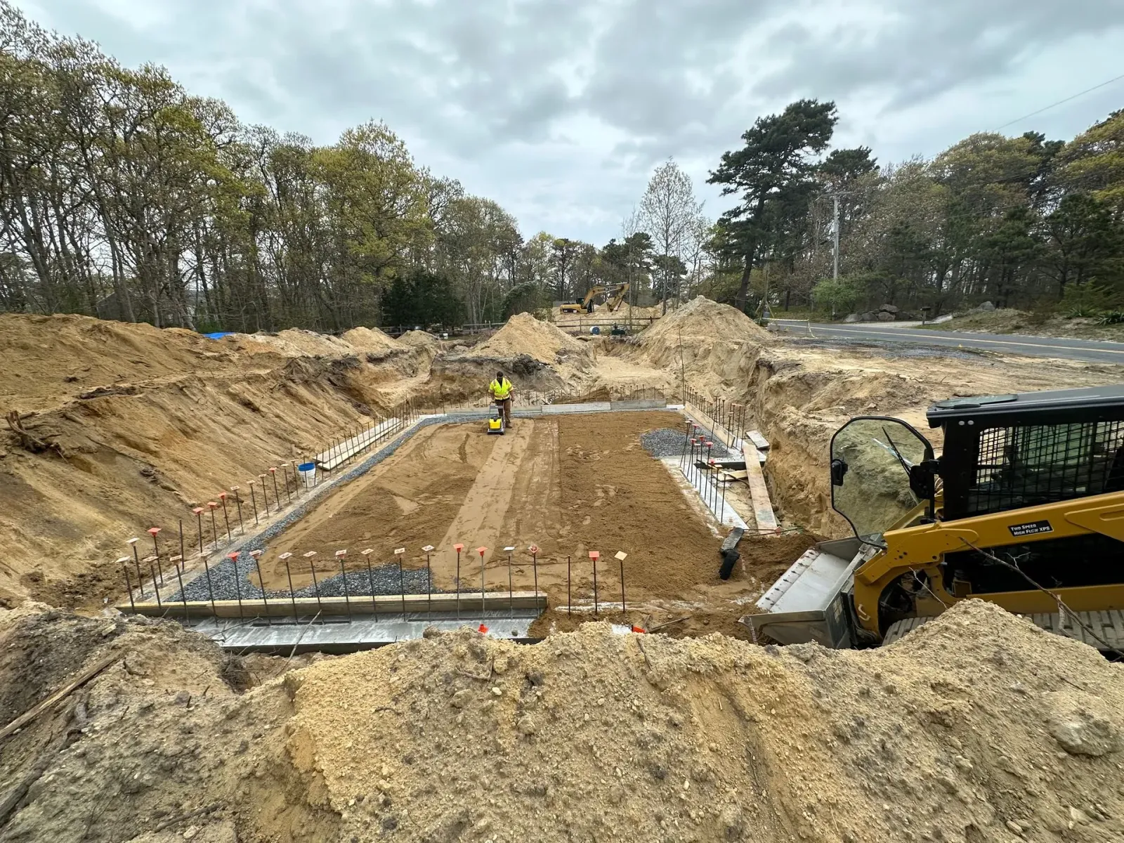 Construction site with excavated earth, concrete forms, and a worker in high-vis vest.