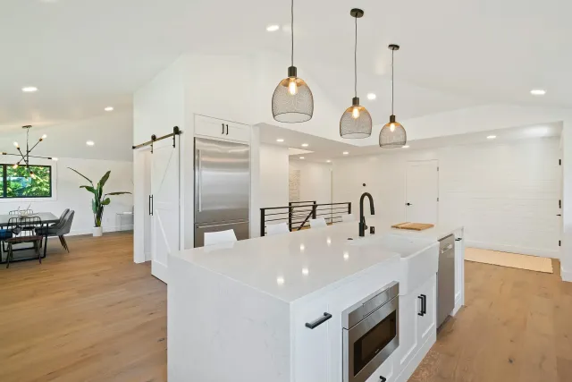 Modern open-concept kitchen with white cabinets, marble countertops, black hardware, pendant lights, and a stainless steel refrigerator, adjacent to a dining area with wooden floor and black chairs.