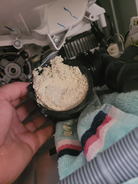 Hand holding a cut-open black pipe filled with white pellets near washing machine parts and a towel