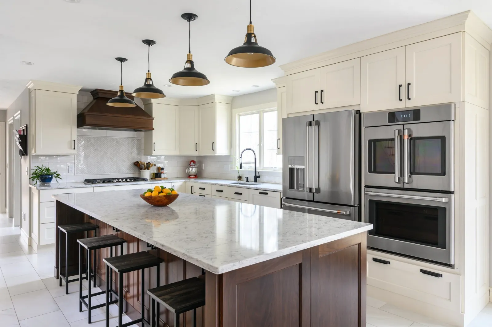Modern kitchen with white cabinets, stainless steel refrigerator, black and gold pendant lighting, and a marble countertop island with three black bar stools and a bowl of fruit.