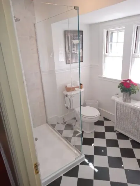 Small bathroom with glass shower, black-and-white checkered floor, toilet, sink, and window.