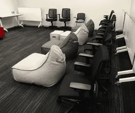 Meeting room with gray bean bags, black chairs, whiteboard, and desks arranged along the walls.
