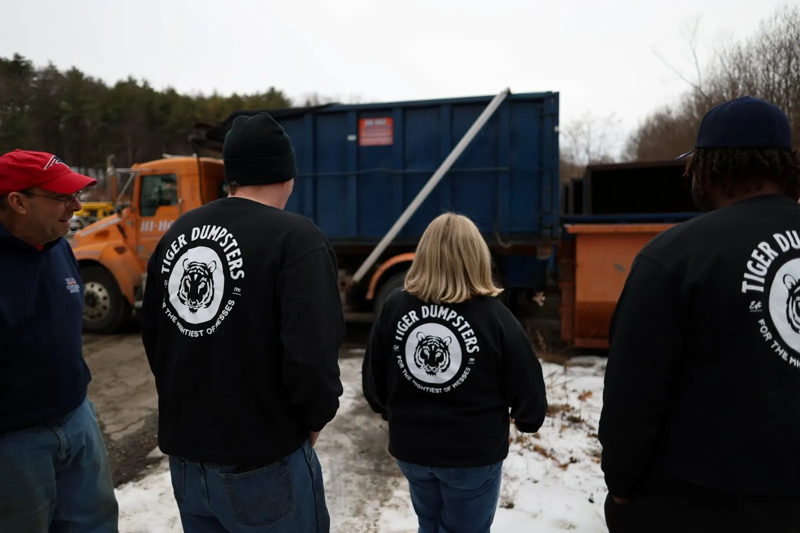 Four people standing outdoors near a blue dump truck on a snowy ground. Three are wearing black jackets with a tiger logo and the words 'Tiger Dumpsters' on the back. The person on the left is an older man in a red cap and navy jacket, talking to the