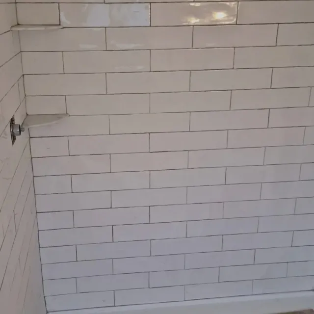 A tiled wall with beige rectangular tiles and a metal pipe outlet on the left side.