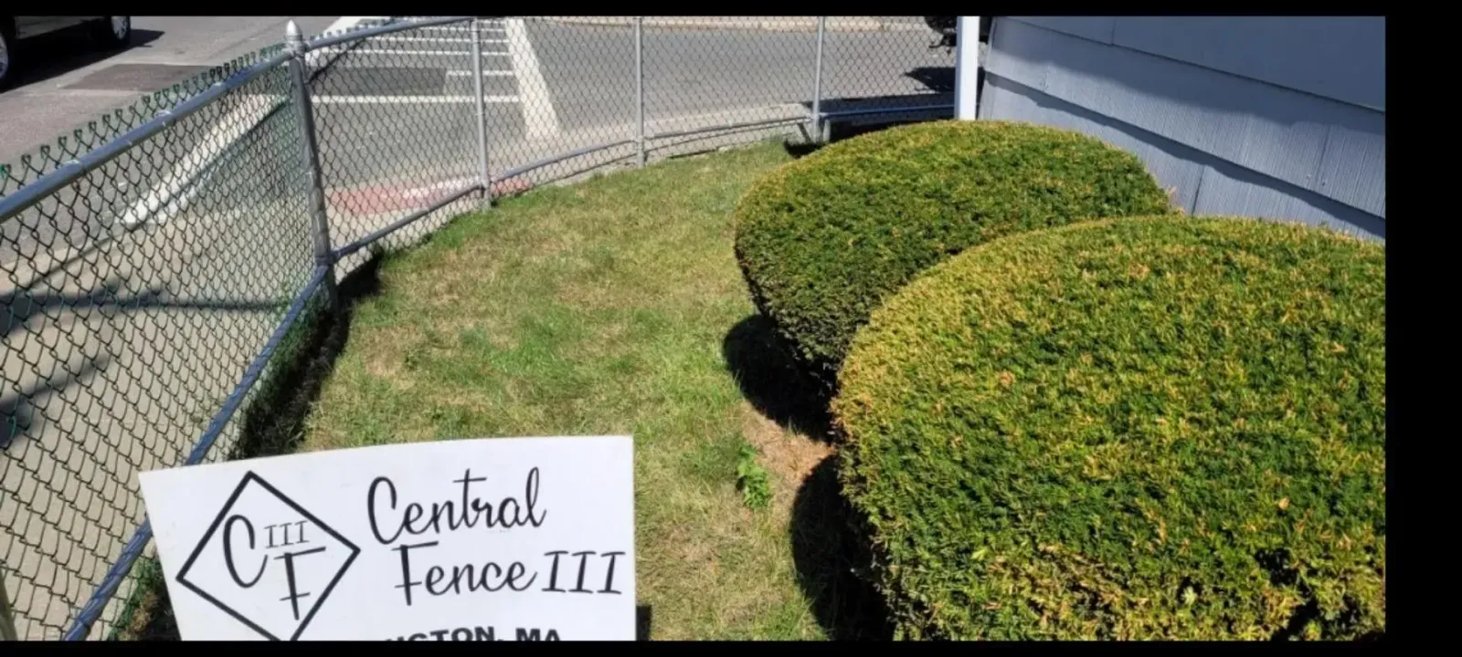 Sign for Central Fence III beside a chain-link fence and neatly trimmed green hedges