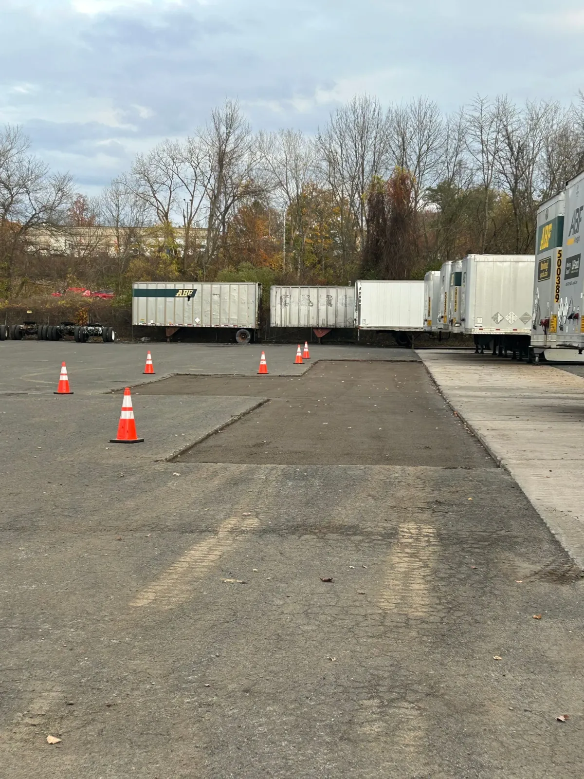 A row of parked white semi-trailers in a gravel lot with orange safety cones demarcating a patch of fresh pavement.