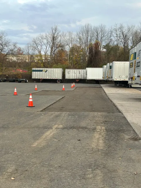 A row of parked white semi-trailers in a gravel lot with orange safety cones demarcating a patch of fresh pavement.