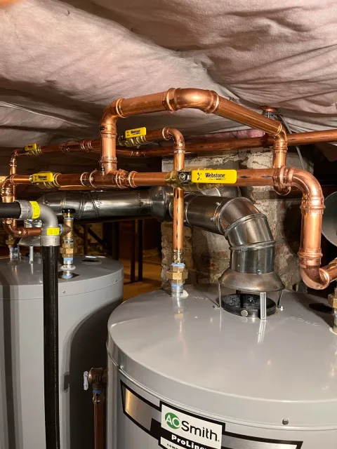 Picture of a plumbing system with copper pipes and valves installed above water heaters, with a white ceiling and brick wall in the background.