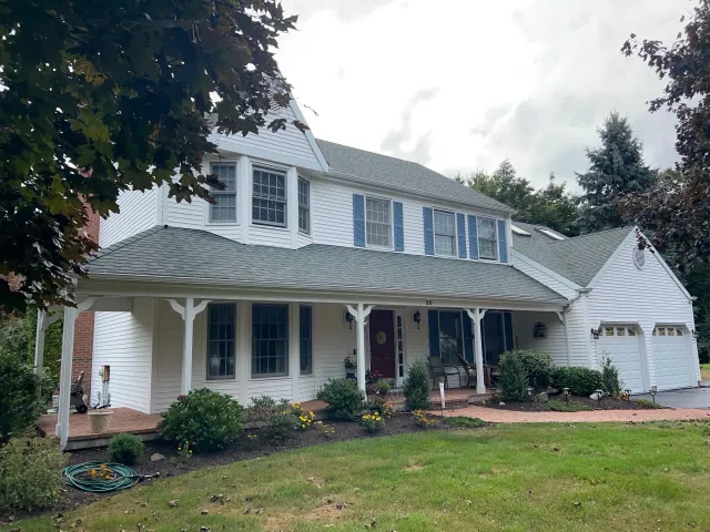 White two-story house with green roof, porch, and attached garage on a grassy lawn under a cloudy sky.