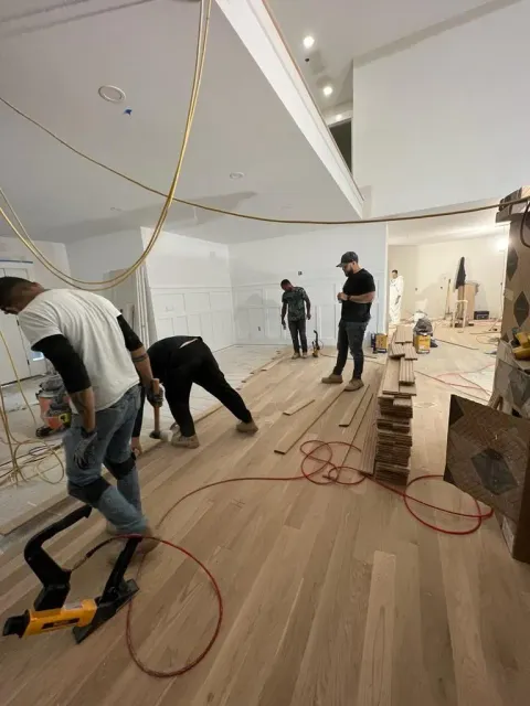 Four people in a partially finished, white-walled room are installing light-colored wood floor planks with tools.