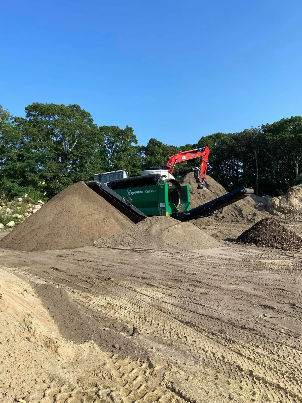 Construction site with soil piles, a green screening machine, and an excavator under a blue sky.