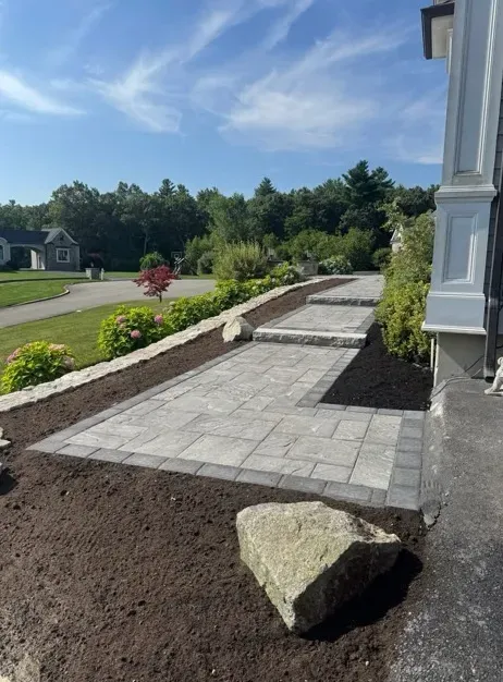 Stone paver walkway with steps, rock border, and dark mulch, leading to a house on a sunny day.