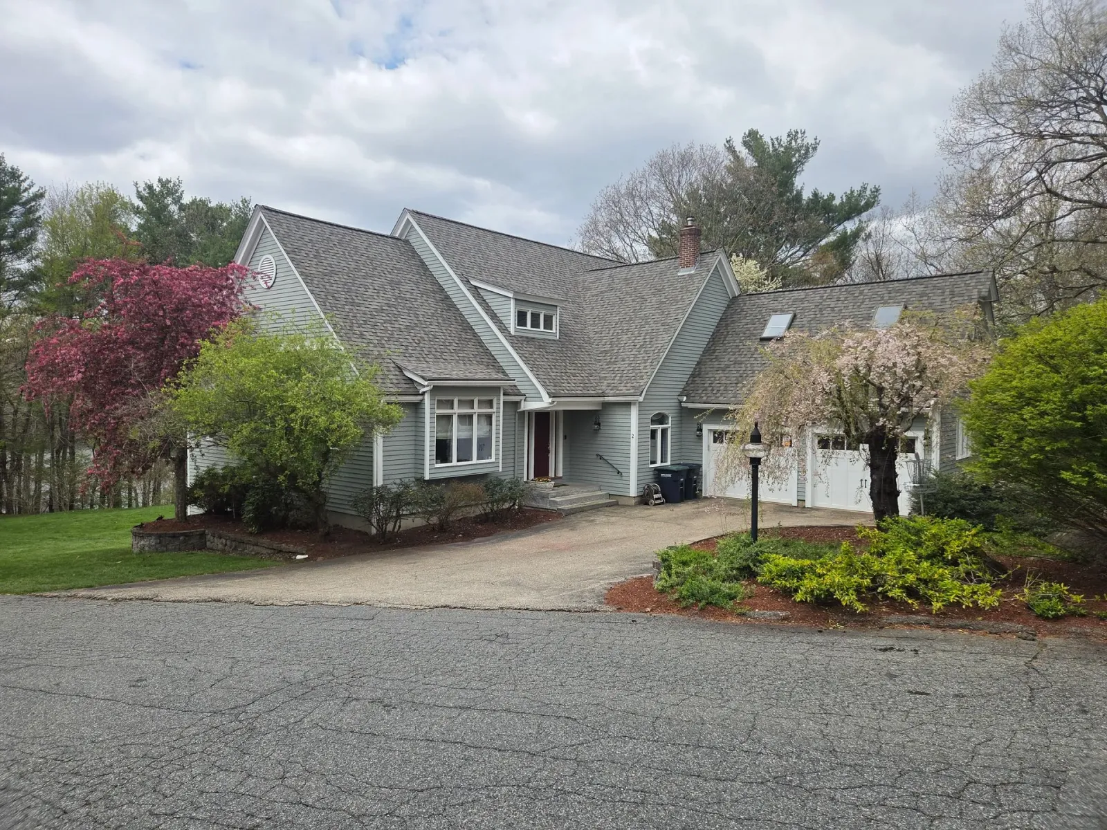 Suburban house with gray roof, front yard, driveway, and spring trees under cloudy sky