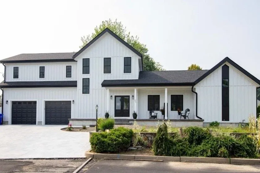 Modern two-story white farmhouse with black trim, featuring a large driveway, two-car garage, covered porch with seating, and minimal landscaping in the front yard.
