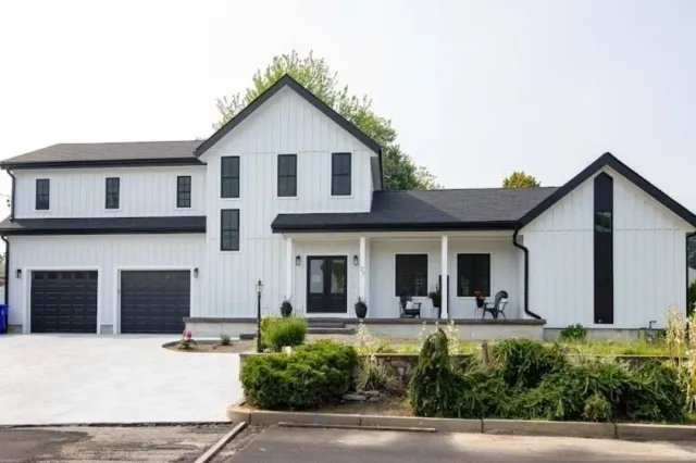 Modern two-story white farmhouse with black trim, featuring a large driveway, two-car garage, covered porch with seating, and minimal landscaping in the front yard.