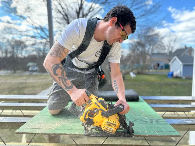 Man with tattoos, wearing safety glasses and black apron, using a yellow DeWalt cordless circular saw to cut a wooden board outdoors on a sunny day.