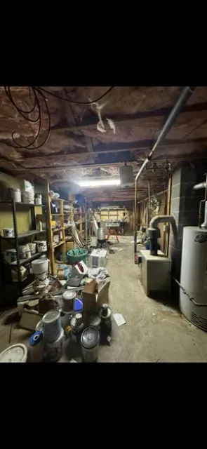 A cluttered, dimly lit basement with exposed ceiling joists, storage shelves, a furnace, and a water heater.