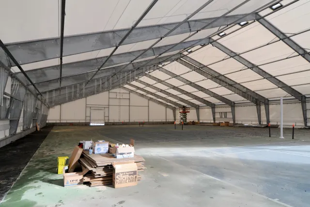 Indoor construction site inside a large tented warehouse with metal framing, scattered boxes and pallets on the floor.