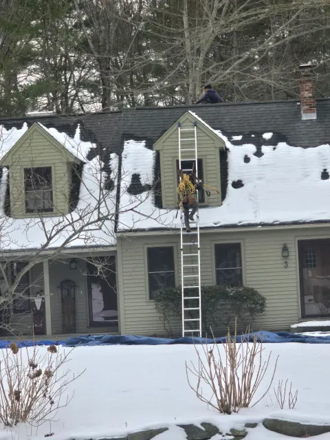 Two-story house with snow-covered roof; person on ladder cleaning an upstairs window.