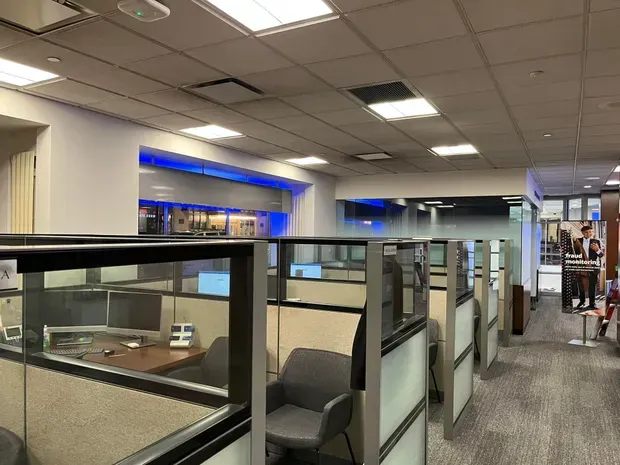 Office cubicles with glass partitions and gray chairs, beneath a drop ceiling with fluorescent lights.