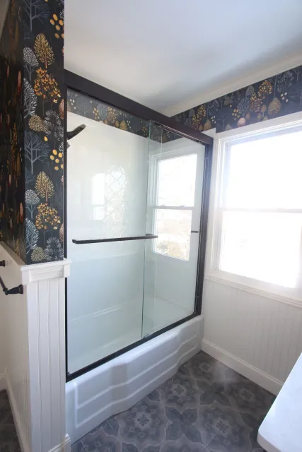 Small bathroom with white tub, glass shower doors, floral wallpaper, and a window.