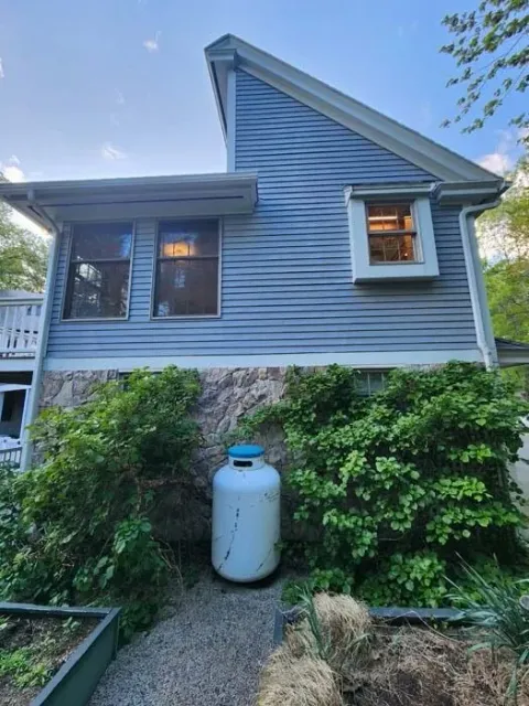 Blue house with a stone foundation, white propane tank, and garden shrubs in front.