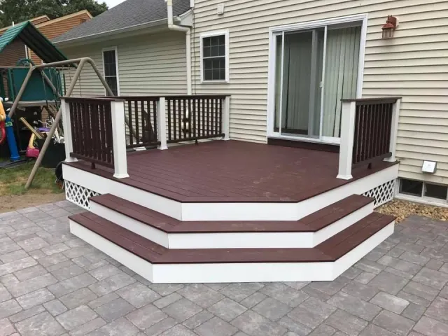 A dark brown composite deck with white railings and two wide steps, built against a tan house with a paved patio.