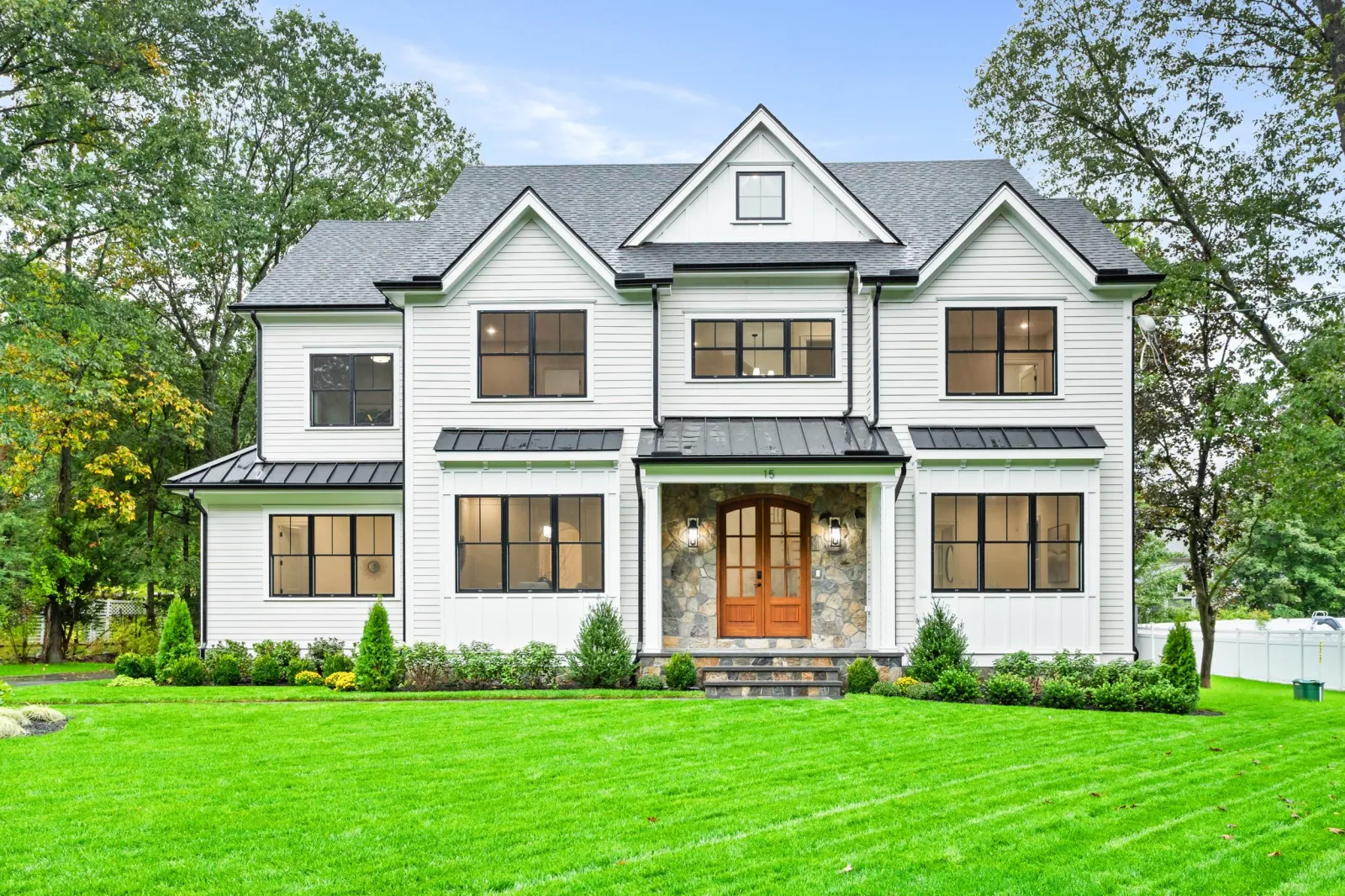 Modern white two-story house with black window frames, dark roof, stone accents on front porch, and well-maintained green lawn with trees in background.
