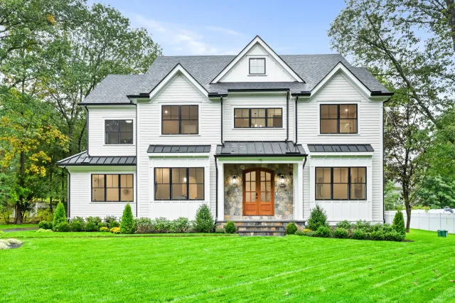 Modern white two-story house with black window frames, dark roof, stone accents on front porch, and well-maintained green lawn with trees in background.