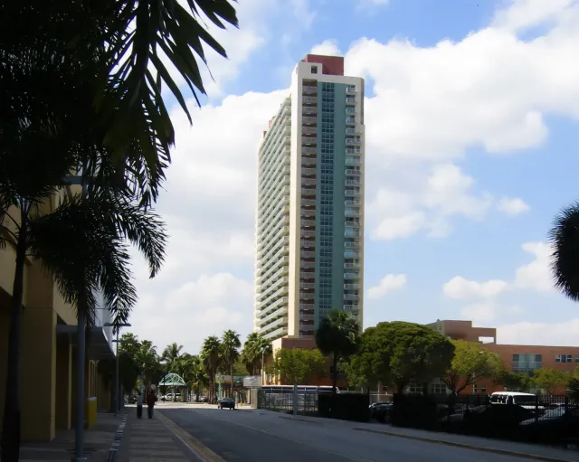 A tall residential building with many balconies and windows stands against a partly cloudy sky. There are palm trees and smaller buildings along a street in the foreground.