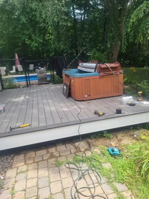 Hot tub on a grey deck with tools scattered nearby. In the background is a pool and trees.