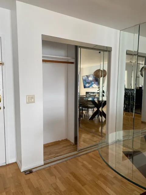 A sliding mirrored closet door partially open in a modern living space with wood flooring and a dining area reflected in the mirror.
