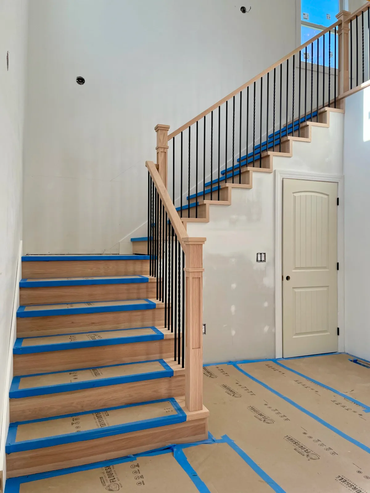 Wooden staircase with black metal balusters, partially finished, in a house under construction with blue painter's tape and unfinished walls.