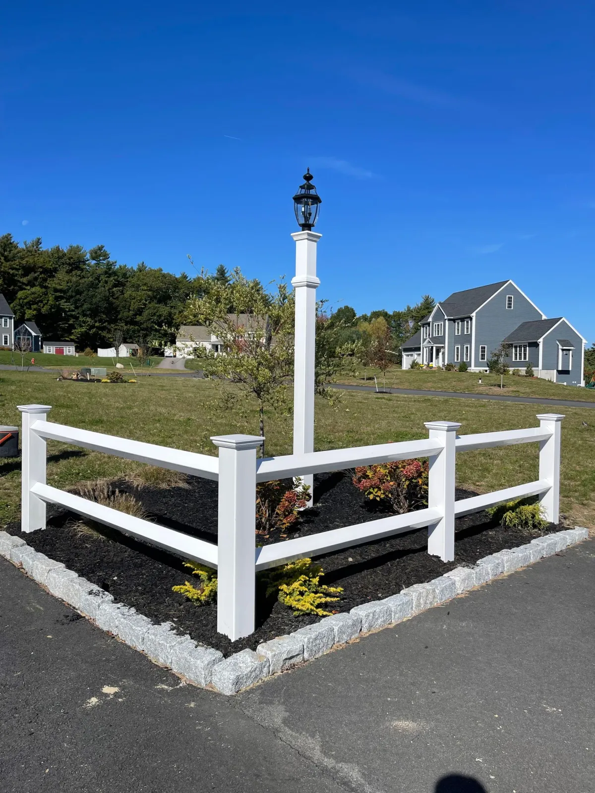 White fence and lamppost on a corner lot with a two-story gray house in the background on a sunny day.
